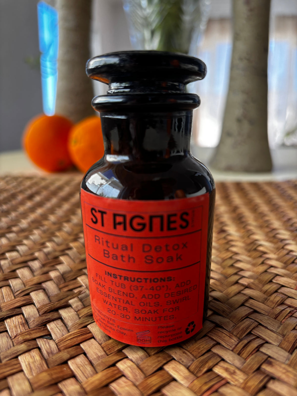 A small dark glass bottle of St Agnes Ritual Detox Bath Soak with a bright orange-red label, placed on a woven wicker surface with a softly blurred indoor background.